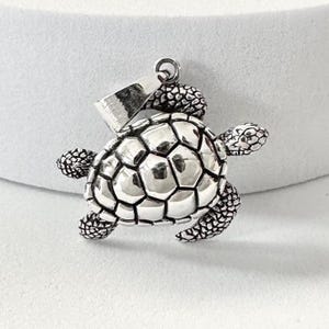 May include: A silver-colored turtle pendant with a detailed shell design. The pendant has a loop at the top for a chain and textured flippers and head. The turtle's shell is made up of geometric shapes.