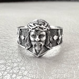Op de afbeelding: Een zilveren ring met een gedetailleerde afbeelding van Jezus die een doornenkroon draagt. De ring heeft een textuur achtergrond met een kruisontwerp aan elke kant.