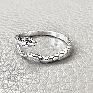 Könnte beinhalten: Ein silberner Ring in Schlangenform. Der Ring zeigt einen detaillierten Schlangenkopf mit geometrischen Mustern und einen Körper, der das Ringband bildet. Der Ring liegt auf einer strukturierten, hellgrauen Oberfläche.