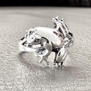 Puede incluir: Anillo plateado con un diseño de rana detallado. La rana se representa en posición sentada, con sus patas y pies palmeados visibles. El anillo tiene un acabado pulido y los rasgos de la rana están claramente definidos.