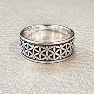 Könnte beinhalten: Ein silberner Ring mit einem sich wiederholenden Blumenmuster um das Band. Der Ring hat eine polierte Oberfläche und ein detailliertes Design. Das Blumenmuster besteht aus miteinander verbundenen Blumenformen und bildet ein durchgehendes Band aus dekorativen Elementen.