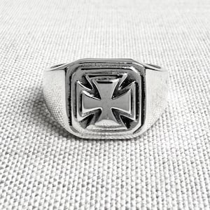 Könnte beinhalten: Ein silberfarbener Ring mit quadratischer Oberfläche, der ein erhabenes Malteserkreuz-Design aufweist. Der Ring hat eine polierte Oberfläche und ein dickes Band. Das Kreuz ist mit abgeschrägten Kanten detailliert, wodurch ein dreidimensionaler Effekt entsteht. Der Ring ist wahrscheinlich aus Metall.