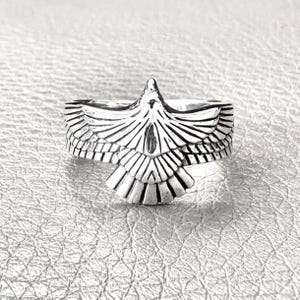 May include: A silver-coloured metal ring featuring a detailed, embossed eagle design with spread wings. The bird motif is centred on the band, with deep, dark oxidised grooves highlighting the individual feathers and the sharp, pointed beak.