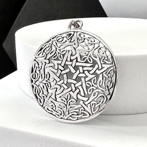 May include: A silver-colored pendant with a Celtic knot design. The circular pendant features an intricate pattern of interwoven lines and shapes, creating a detailed and textured surface. The pendant has a small loop at the top for a chain.