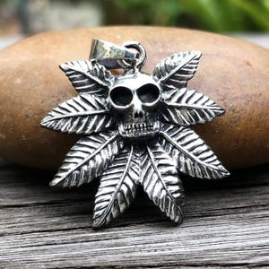 Puede incluir: Colgante de plata con un diseño de calavera y hoja de marihuana. El colgante es detallado e intrincado.