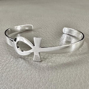 Può includere: Un braccialetto d'argento con un simbolo Ankh. Il braccialetto è realizzato in metallo liscio e lucido e ha un design semplice ed elegante.