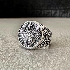 May include: A silver ring with a detailed design featuring an angel with wings holding a sword and shield. The ring has a round face and a textured band.