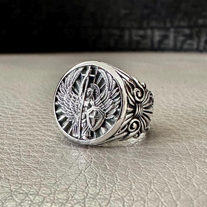 May include: A silver ring with a detailed design featuring an angel with wings holding a sword and shield. The ring has a round face and a textured band.