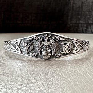 Op de afbeelding: Zilverkleurige manchetarmband met ingewikkelde reliëfdesigns. Voorzien van een centrale gevleugelde figuur met een schedel, geflankeerd door andere symbolen en Keltische knoopwerk. De armband heeft een donkere achtergrond.