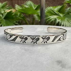 May include: A silver cuff bracelet with a Native American design. The bracelet features Kokopelli figures and geometric patterns. The open cuff design allows for easy wear. The bracelet is displayed on a light grey surface with green foliage in the background.