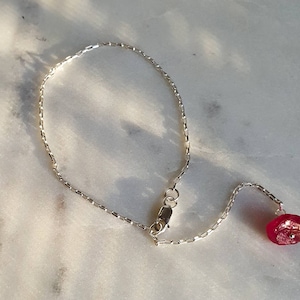 May include: A silver chain bracelet with a red gemstone pendant. The chain is a delicate box chain style.