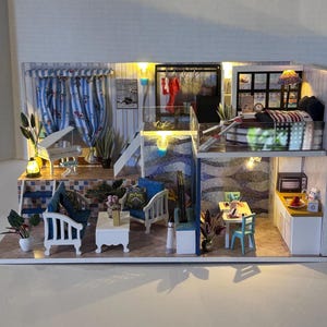 Könnte beinhalten: Ein Miniatur-Puppenhaus mit zwei Etagen, mit einem Wohnzimmer mit einem weißen Sofa, einem Couchtisch und einem blauen Sessel, einer Küche mit einer weißen Arbeitsplatte und einer Mikrowelle sowie einem Schlafzimmer mit einem Bett, einem Nachttisch und einer Lampe. Das Puppenhaus ist mit blau-weiß gemusterter Tapete dekoriert, und es gibt überall Pflanzen und Blumen.