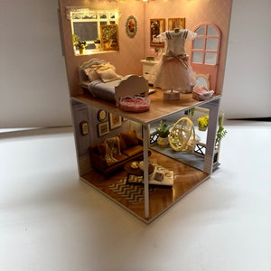 May include: A miniature dollhouse with two levels, featuring a bedroom with a bed, a living room with a sofa and a coffee table, and a balcony with a swing. The dollhouse is decorated with furniture, plants, and artwork.