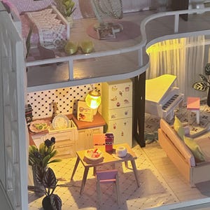Puede incluir: Una casa de muñecas en miniatura con múltiples habitaciones, incluyendo una cocina, sala de estar y dormitorio. La casa de muñecas presenta un piano blanco, una pequeña mesa con comida y plantas decorativas. La iluminación crea un ambiente cálido.