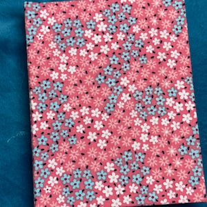 Puede incluir: Un cuaderno rosa con un patrón floral con flores blancas y azules. El cuaderno tiene una cubierta suave y texturizada.