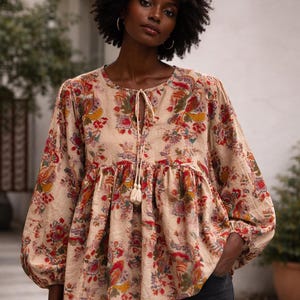 May include: A beige floral print blouse with long sleeves and a tie-neck detail. The blouse features a round neckline and a gathered waist. The floral pattern includes red, orange, and green accents. The model is wearing black jeans.