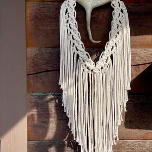 May include: A cream-colored macrame wall hanging with a natural antler base. The macrame features a woven top section and long, fringed strands. The piece is suspended against a wooden background, creating a rustic, bohemian aesthetic.