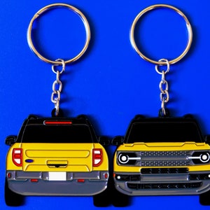 Peut inclure: Deux porte-clés Ford Bronco jaunes avec des anneaux de clés argentés. Les porte-clés sont en métal et présentent une vue avant et arrière du véhicule.
