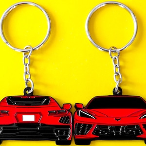 May include: Two metal keychains featuring red sports car designs. One keychain shows the rear view of the vehicle with black detailing and a white license plate area, while the other shows the front view with black headlights and grille accents.