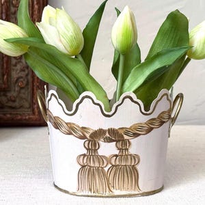 May include: A white metal planter with a scalloped top edge and two side handles, featuring a painted gold rope and tassel design around the exterior. The planter is filled with several white and green tulip flowers with long green leaves.