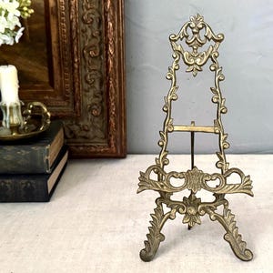 May include: A brass display easel featuring an intricate, ornate metalwork design with scrolling patterns, a central support bar, and a wide base. The stand has a metallic gold-toned finish and a tall, pointed structure for holding items.