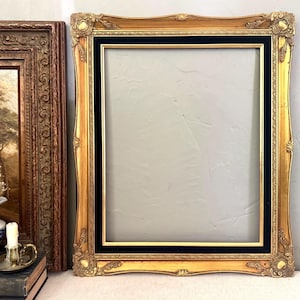 May include: Ornate gold picture frame with a black inner border. The frame has detailed carvings and a rectangular opening. A second, smaller framed artwork is visible to the left.