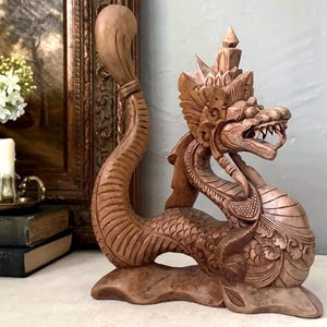 May include: A light brown wooden sculpture of a mythical dragon creature with an elongated, curved tail, intricate scale carvings, and a detailed head featuring an open mouth with visible teeth and a pointed, ornate crown-like crest on its head.