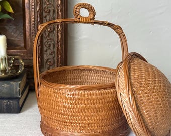 May include: A round, light brown woven wicker basket with a matching domed lid and a tall, arched handle featuring a small decorative loop at the top. The basket has a tight, intricate weave pattern and a sturdy base, presented with the lid set aside.