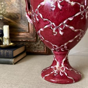 May include: A deep red ceramic urn featuring a glossy finish and a decorative pattern of white ribbons tied into bows that drape across the surface. The vessel has a pedestal base and two side handles, showcasing a vibrant, solid color scheme.