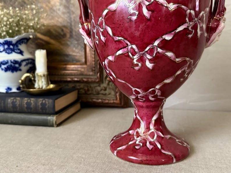 May include: A deep red ceramic urn featuring a glossy finish and a decorative pattern of white ribbons tied into bows that drape across the surface. The vessel has a pedestal base and two side handles, showcasing a vibrant, solid color scheme.