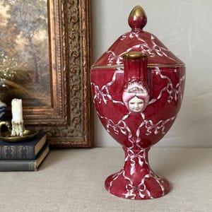May include: A deep red ceramic lidded urn featuring a white ribbon pattern and a sculpted face detail on the handle. The vessel has a pedestal base and a pointed lid with gold-toned accents on the finial and handle top, standing upright.