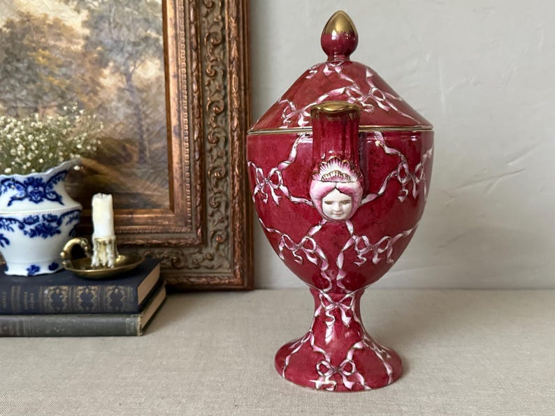 May include: A deep red ceramic lidded urn featuring a white ribbon pattern and a sculpted face detail on the handle. The vessel has a pedestal base and a pointed lid with gold-toned accents on the finial and handle top, standing upright.