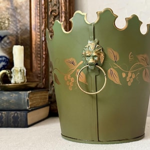 May include: An olive green metal bucket with a scalloped top edge, featuring a gold-toned lion head ornament holding a circular ring handle. The exterior is decorated with painted gold leaf and vine motifs wrapping around the cylindrical body.