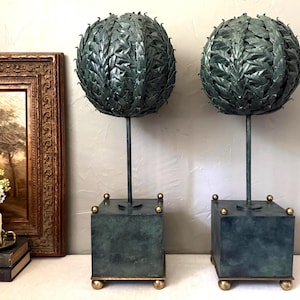 May include: A pair of decorative sculptures featuring spherical tops composed of layered, dark green metal leaves. Each sphere is supported by a thin vertical rod attached to a square, dark green base with small gold-toned spherical feet and accents.
