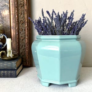 May include: A light blue, hexagonal ceramic planter filled with dried lavender. The glossy vase has a geometric design and sits on a white surface. A vintage painting and other decorative items are in the background.