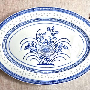 May include: An oval ceramic serving platter featuring a white base with blue floral illustrations in the center. The rim has a decorative pierced lattice pattern and a blue geometric border. The design includes a central sunflower-like motif.