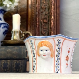 May include: A white ceramic bowl with a scalloped blue rim, featuring a relief-molded face with orange hair on the front. The exterior is decorated with vertical orange stripes, blue and green vine patterns, and small orange floral accents.