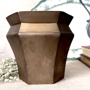May include: A bronze-colored, geometric planter with a wide, octagonal shape. The planter has a slightly flared top and a textured surface. It is displayed with dried white flowers, books, and a decorative metal object.