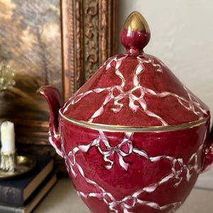 May include: A deep red ceramic sugar bowl with a matching lid, featuring a white and pink ribbon and bow pattern across the surface. The bowl has two curved handles and a gold-toned finial on the lid, with gold trim along the rim and handles.