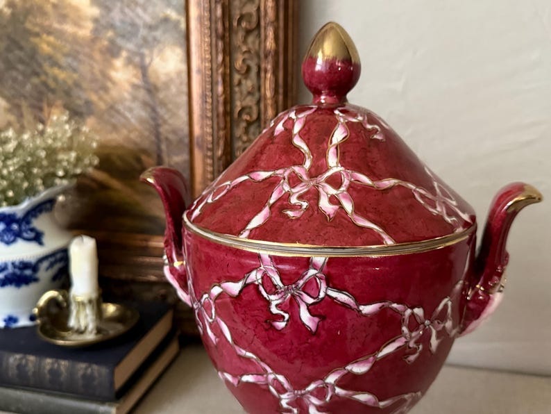 May include: A deep red ceramic sugar bowl with a matching lid, featuring a white and pink ribbon and bow pattern across the surface. The bowl has two curved handles and a gold-toned finial on the lid, with gold trim along the rim and handles.