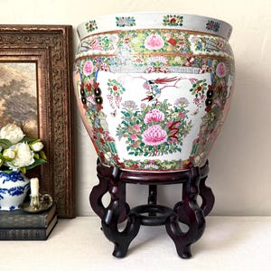 May include: A large, round, white ceramic fishbowl planter featuring colorful hand-painted floral patterns, butterflies, and birds. The vessel sits on a dark brown, carved wooden stand with four curved legs and a circular base support.