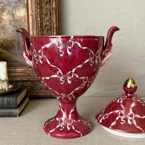 May include: A deep red ceramic lidded urn featuring a white ribbon and bow pattern across the exterior. The urn has two ornate handles on the sides and a matching removable lid with a gold-toned finial. The interior rim is trimmed in gold.
