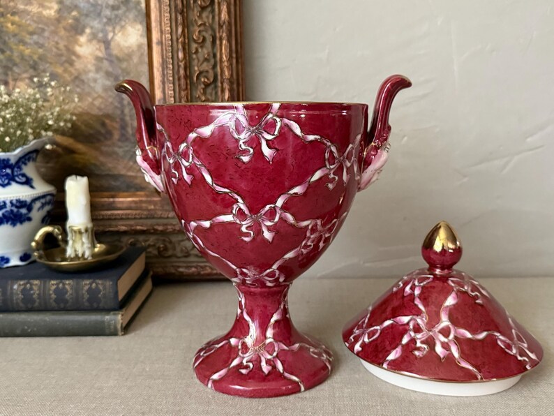 May include: A deep red ceramic lidded urn featuring a white ribbon and bow pattern across the exterior. The urn has two ornate handles on the sides and a matching removable lid with a gold-toned finial. The interior rim is trimmed in gold.