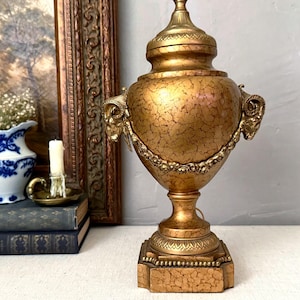 May include: A gold-colored, ornate table lamp with a textured finish. The lamp features ram's head accents and a decorative garland. The base is square and textured. A blue and white vase, books, and a candle are in the background.