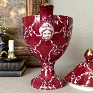 May include: A deep red ceramic chalice-shaped vessel with a lid, featuring a raised white ribbon pattern and a central sculpted face with a gold-trimmed top. The piece has a pedestal base and gold accents on the rim and the finial of the lid.