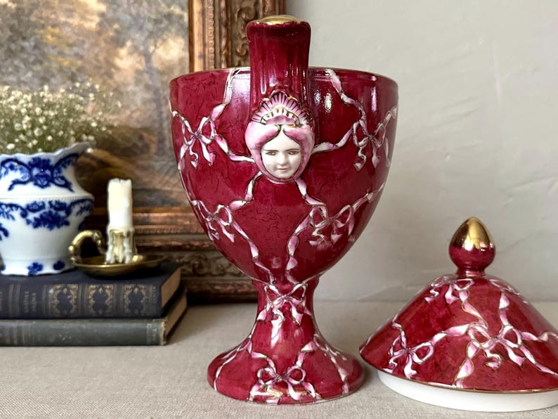 May include: A deep red ceramic chalice-shaped vessel with a lid, featuring a raised white ribbon pattern and a central sculpted face with a gold-trimmed top. The piece has a pedestal base and gold accents on the rim and the finial of the lid.