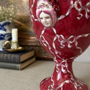 May include: A deep red ceramic urn featuring a raised, three-dimensional porcelain face of a person wearing a bonnet. The surface is decorated with painted white ribbons and bows outlined in gold trim, extending from the base to the rim.