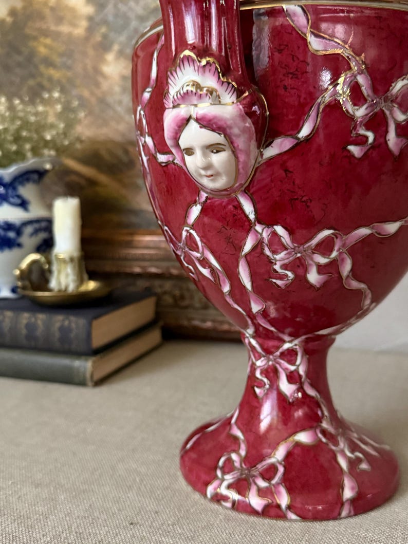 May include: A deep red ceramic urn featuring a raised, three-dimensional porcelain face of a person wearing a bonnet. The surface is decorated with painted white ribbons and bows outlined in gold trim, extending from the base to the rim.