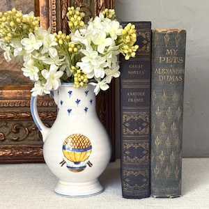 May include: A white ceramic pitcher with a blue handle and small blue floral accents, featuring a yellow and brown hot air balloon illustration. The pitcher is filled with white and yellow flowers and stands next to two vintage books with gold text.