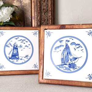 May include: Two square ceramic tiles framed in natural wood. Each tile features a white background with a blue circular border containing a detailed blue illustration. One tile depicts a windmill, while the other shows a sailboat on water.